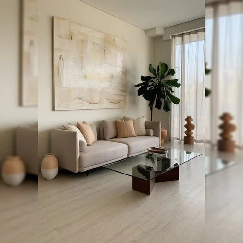 A photo of a modern living room with a couch and a glass coffee table. There is a potted plant in the corner. The wall has a large, abstract painting. The floor has a light wooden hue