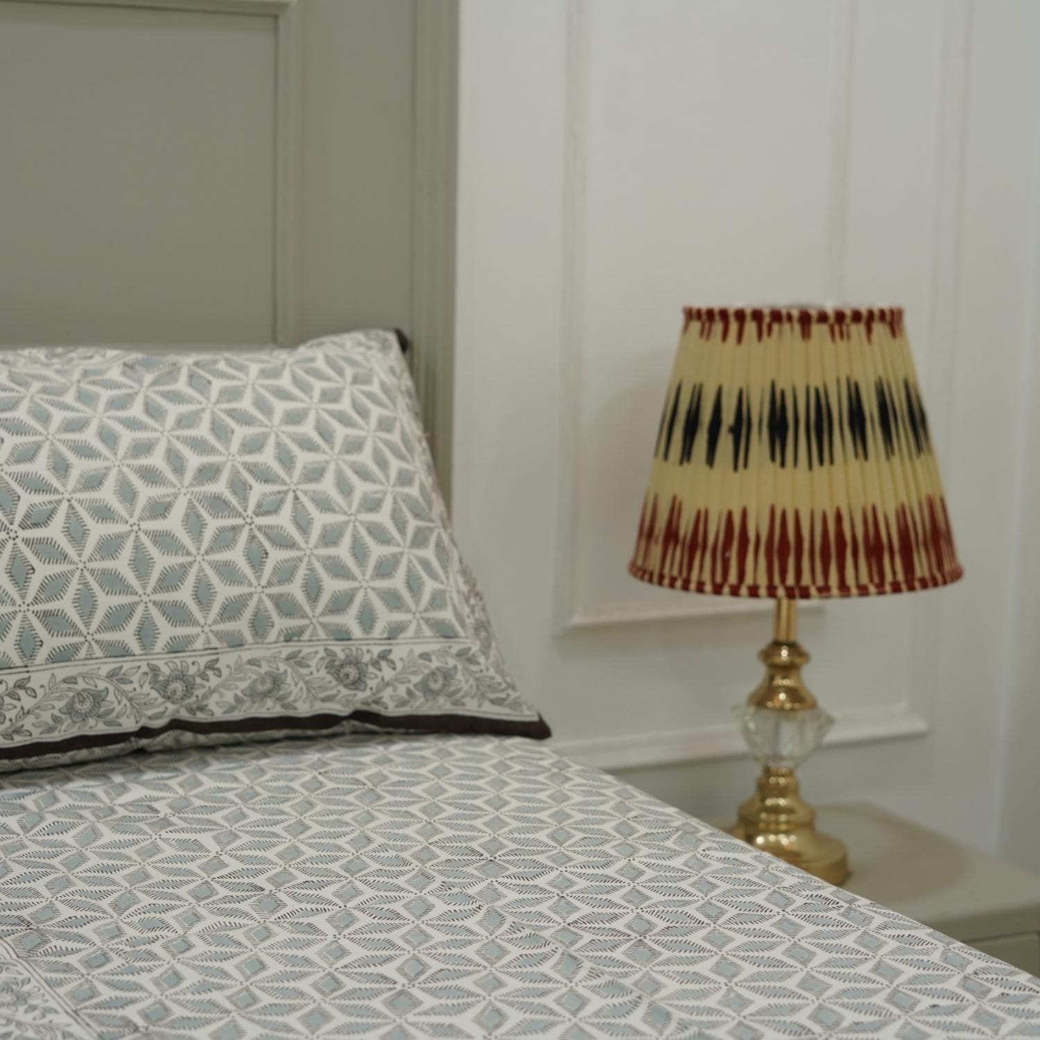 Bed with patterned bedding and pillow, nightstand with lamp against a white wall.