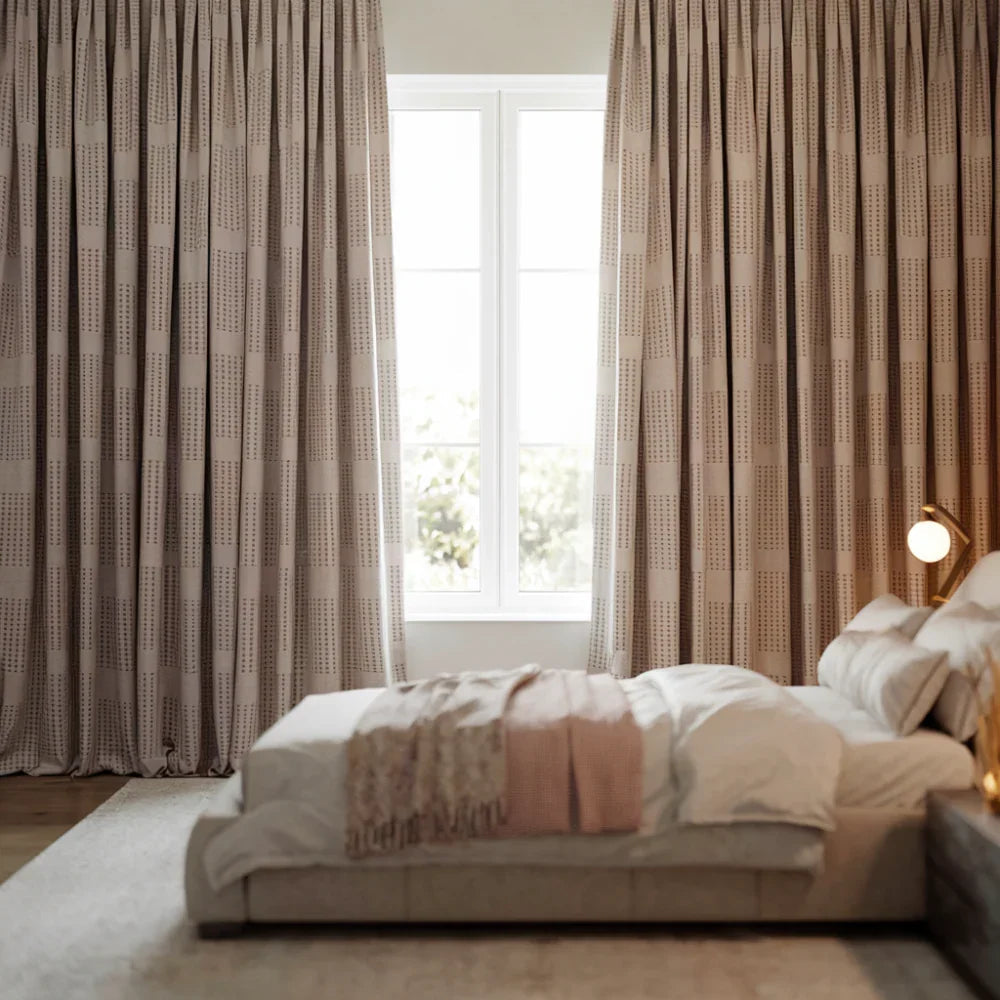 A photo of a serene bedroom with floor-to-ceiling blackout block print curtains in soft neutral tones. The curtains frame a large window, creating a cozy and private retreat.