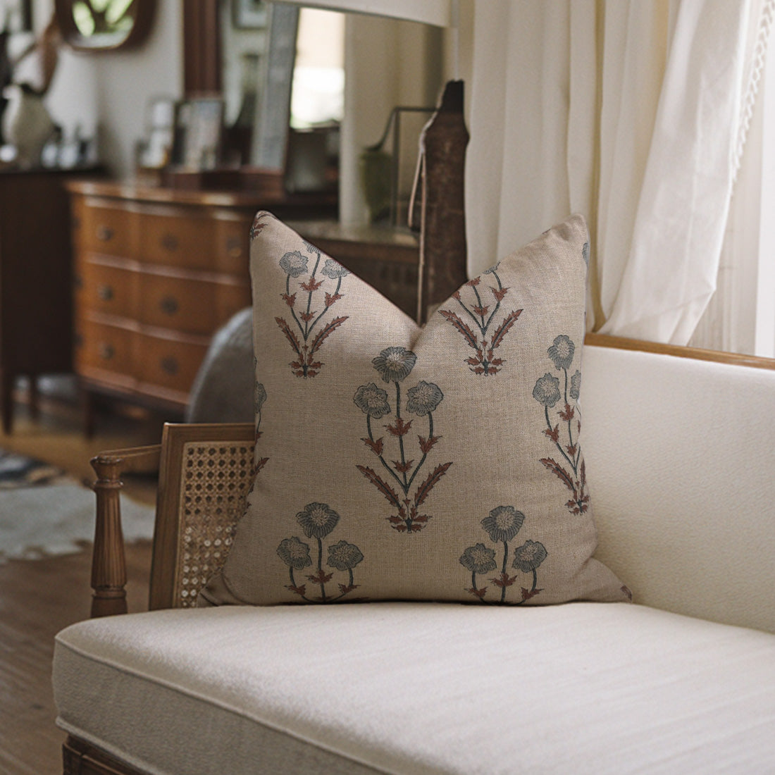 Decorative pillow with a floral pattern on a white sofa in a room with wooden furniture and curtains, styled with decorative cushion covers.